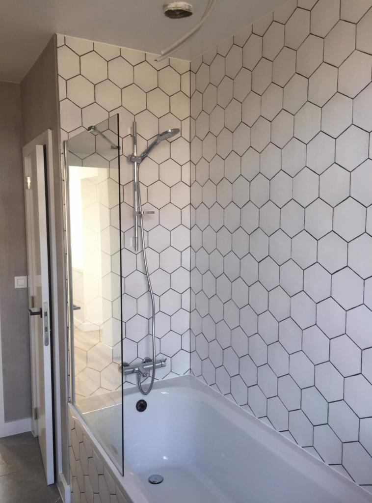 Biała wanna z parawanem i chromowaną baterią, ściana wyłożona białymi heksagonalnymi płytkami z czarnym obrysem, widok na wnętrze łazienki.