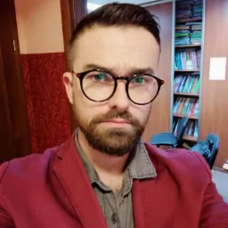 Portret mężczyzny w okularach i bordowej marynarce na tle regału z książkami.