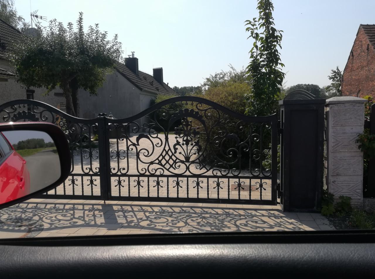Kuta brama wjazdowa z ozdobnymi detalami, widoczna z wnętrza samochodu. Cień bramy na jasnej kostce brukowej. W tle budynki mieszkalne i zieleń.