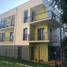 Nowoczesny, trzypiętrowy budynek mieszkalny z balkonami o stalowych balustradach, pomalowany w odcieniach beżu i oliwki, ogrodzony siatką.