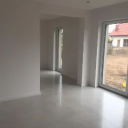 Jasne, przestronne wnętrze domu z białymi ścianami, błyszczącą podłogą z płytek i dużymi oknami przesuwnymi z widokiem na ogród w trakcie przygotowania.