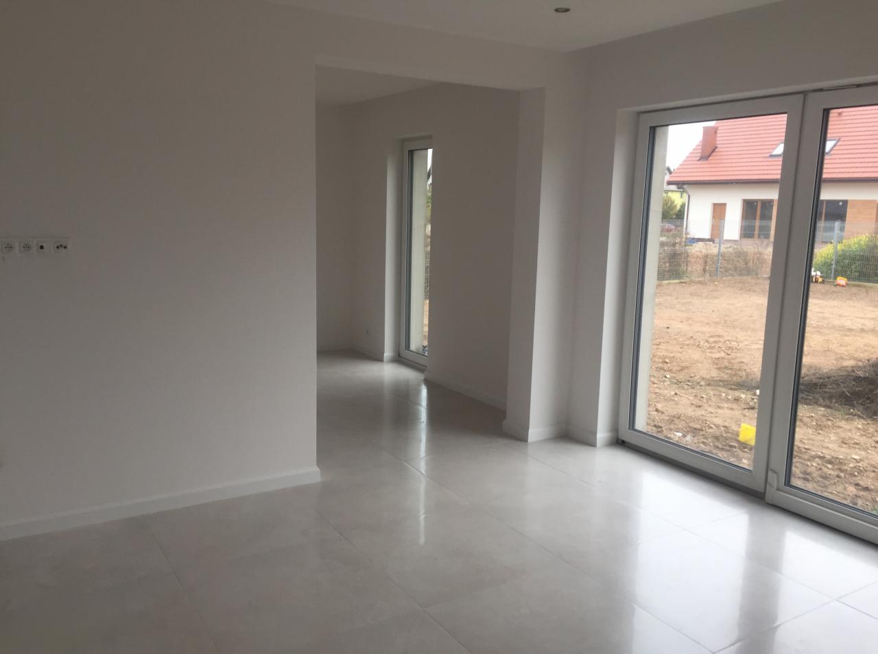 Jasne, przestronne wnętrze domu z białymi ścianami, błyszczącą podłogą z płytek i dużymi oknami przesuwnymi z widokiem na ogród w trakcie przygotowania.