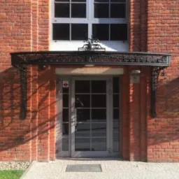 Wejście do budynku z cegły z metalowym daszkiem z greckim wzorem, szare drzwi z szybami i chodnik z kostki.