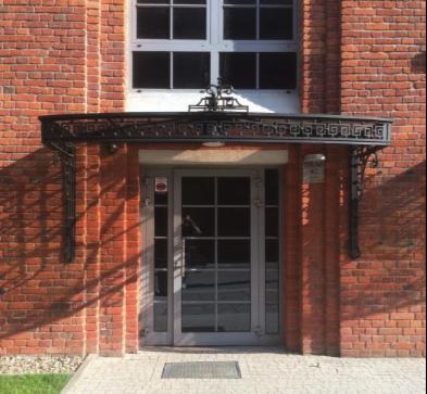 Wejście do budynku z cegły z metalowym daszkiem z greckim wzorem, szare drzwi z szybami i chodnik z kostki.