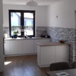 Jasna kuchnia z białymi szafkami, drewnianym blatem i panelami imitującymi kamień na ścianie, widok na zewnątrz przez czarne okno, drewnopodobne płytki na podłodze i stół z krzesłami w jadalni.