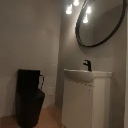 Nowoczesna łazienka z czarną toaletą i owalnym lustrem. Biała szafka pod umywalką z czarną armaturą, oświetlenie punktowe. Jasne płytki na podłodze i ścianach.