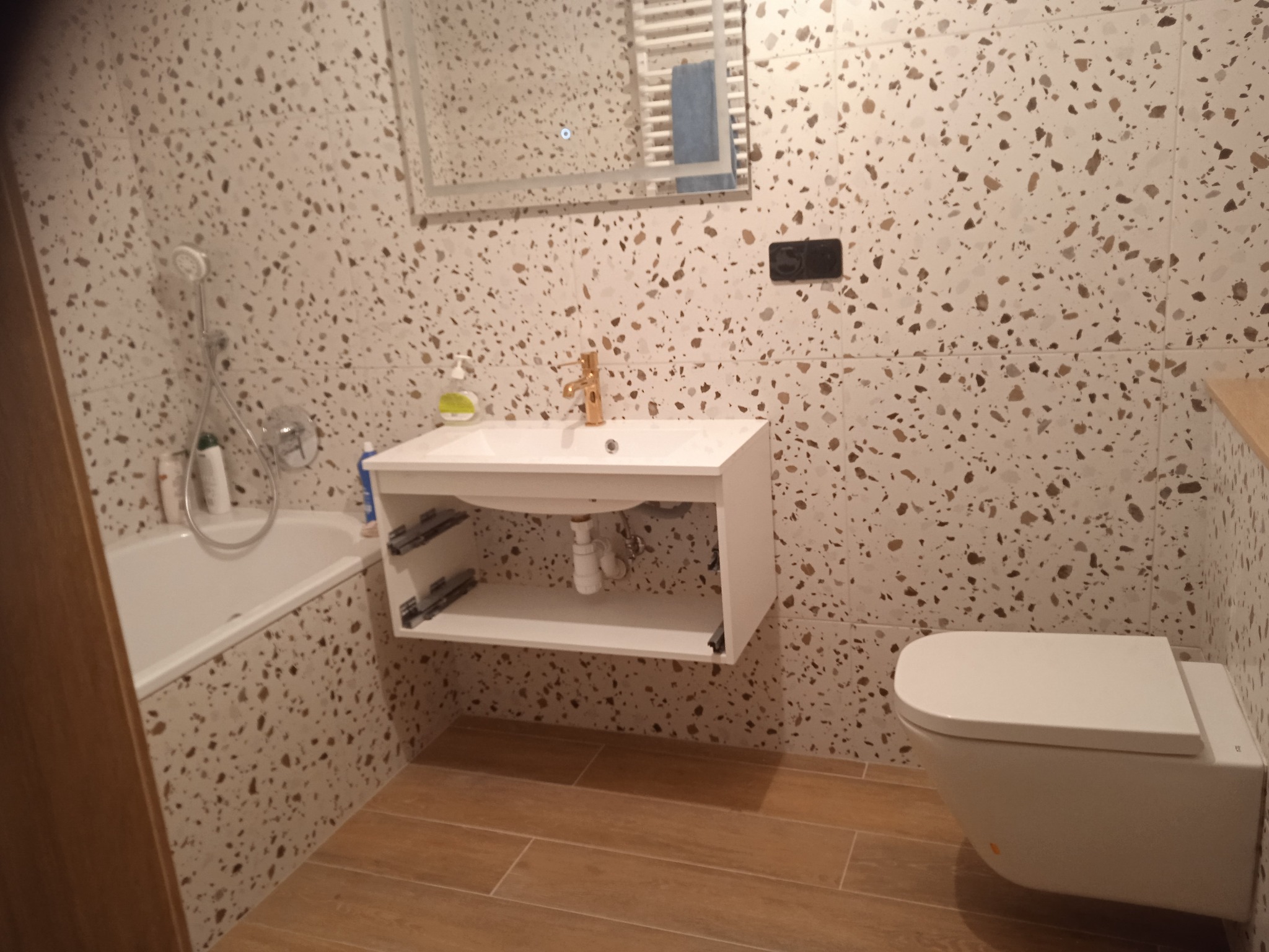 Wykończona łazienka z armaturą w kolorze złota, płytkami lastryko na ścianach i podłodze imitującą drewno. Widoczna umywalka z szafką, toaleta i wanna.