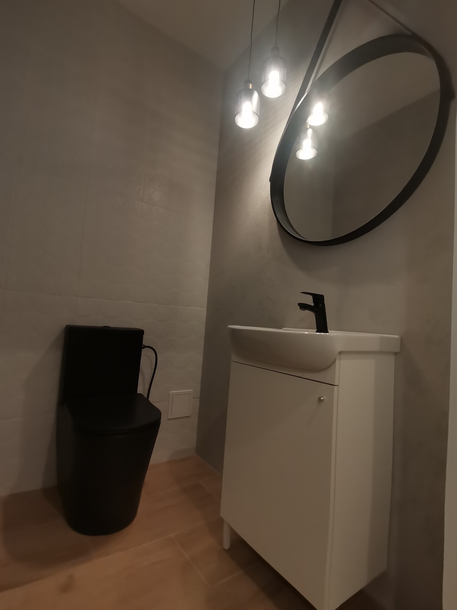 Nowoczesna łazienka z czarną toaletą i owalnym lustrem. Biała szafka pod umywalką z czarną armaturą, oświetlenie punktowe. Jasne płytki na podłodze i ścianach.