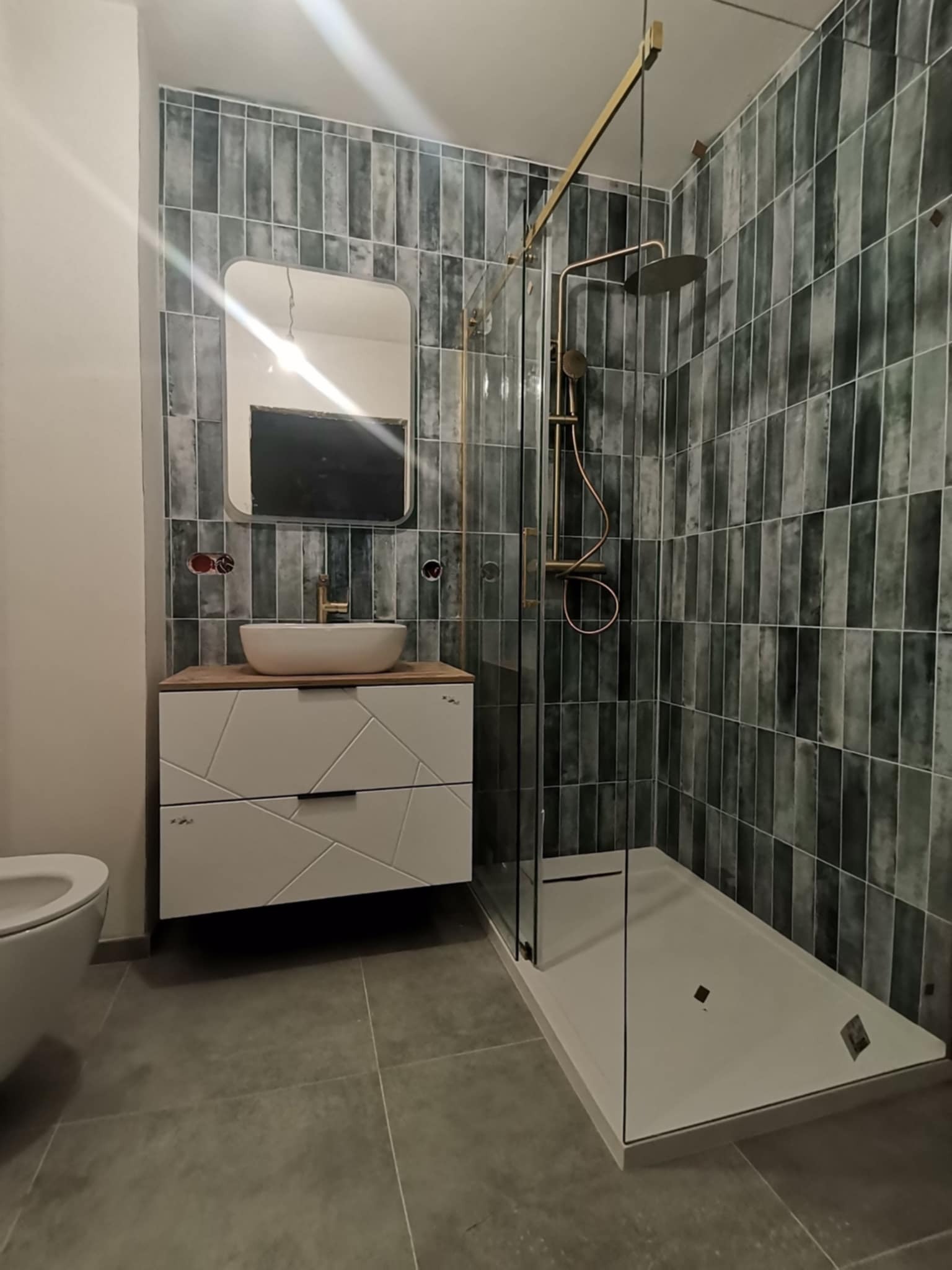 Nowoczesna łazienka po remoncie: kabina prysznicowa ze złotymi elementami, umywalka nablatowa na szafce z geometrycznym wzorem, płytki w odcieniach szarości i zieleni.