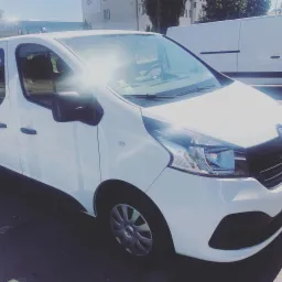 Biały Renault Trafic, widok z przodu, z silnym odblaskiem słońca na przedniej szybie, zaparkowany na zewnątrz w słoneczny dzień.