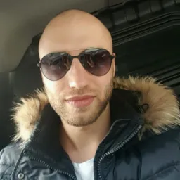 Mężczyzna w kurtce z futrzanym kapturem i okularach przeciwsłonecznych siedzi w samochodzie. Widoczna tapicerka i uchwyt podsufitki.