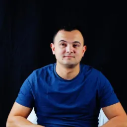 Portret mężczyzny w niebieskiej koszulce na czarnym tle, z delikatnym uśmiechem i pieprzykiem na policzku.