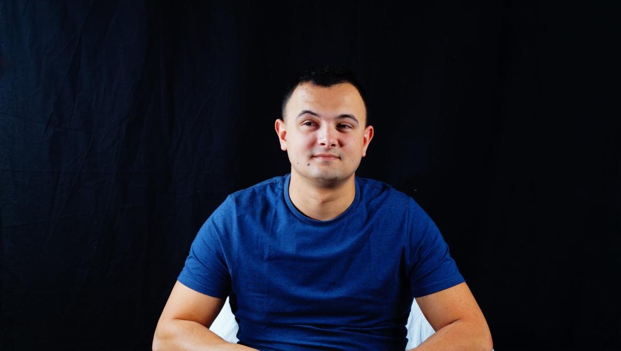 Portret mężczyzny w niebieskiej koszulce na czarnym tle, z delikatnym uśmiechem i pieprzykiem na policzku.