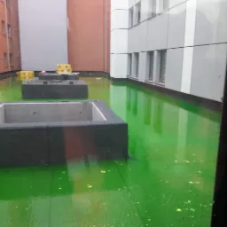 Zielona, płaska powierzchnia dachu pokryta płynną substancją, prawdopodobnie po impregnacji lub czyszczeniu, z widocznymi elementami konstrukcyjnymi i budynkami w tle.