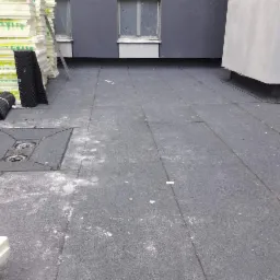 Płaski dach pokryty papą z widocznymi elementami wentylacji, izolacją termiczną XPS i fragmentem drabiny opartą o ścianę budynku z oknami.