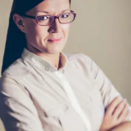 Portret kobiety w okularach, z założonymi rękami, w jasnej koszuli. Uśmiechnięta, z delikatnym makijażem, na neutralnym tle. Profesjonalny wizerunek.