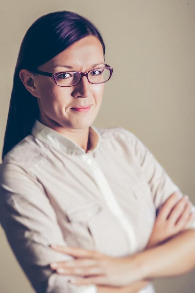 Portret kobiety w okularach, z założonymi rękami, w jasnej koszuli. Uśmiechnięta, z delikatnym makijażem, na neutralnym tle. Profesjonalny wizerunek.