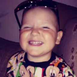 Uśmiechnięty chłopiec w koszulce z motywem Myszki Miki, z okularami przeciwsłonecznymi na głowie, przymyka oczy w radosnym grymasie.