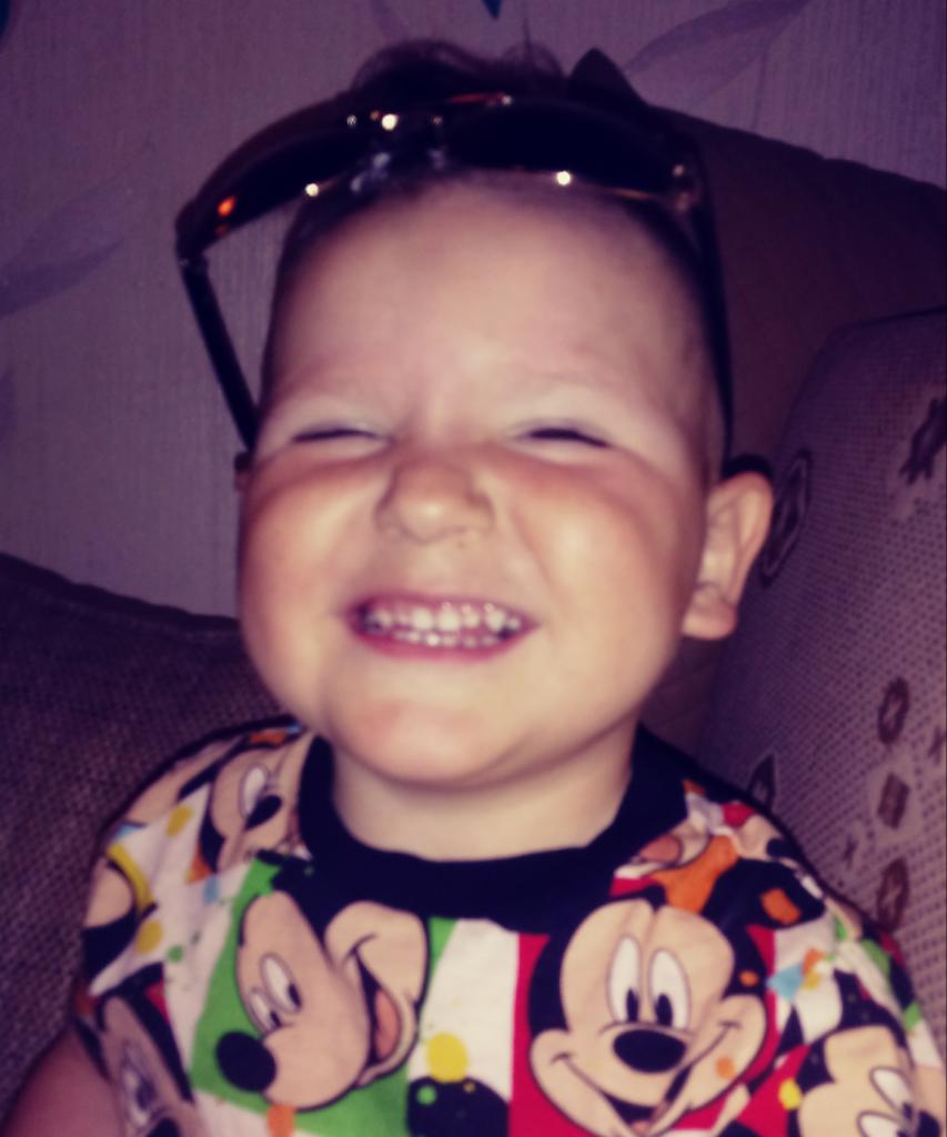 Uśmiechnięty chłopiec w koszulce z motywem Myszki Miki, z okularami przeciwsłonecznymi na głowie, przymyka oczy w radosnym grymasie.