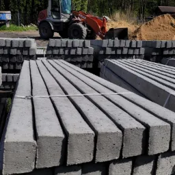 Stosy szarych, kwadratowych słupków betonowych na paletach, z koparką Atlas i stertą piasku w tle.