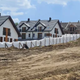 Nowe domy jednorodzinne z szarymi dachami i białymi elewacjami, oddzielone płotem z siatki budowlanej od placu budowy z widoczną ziemią i śladami opon.