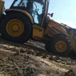 Żółta koparko-ładowarka Komatsu na nierównym, piaszczystym terenie w trakcie prac ziemnych, widoczne świeże ślady po wykopie.