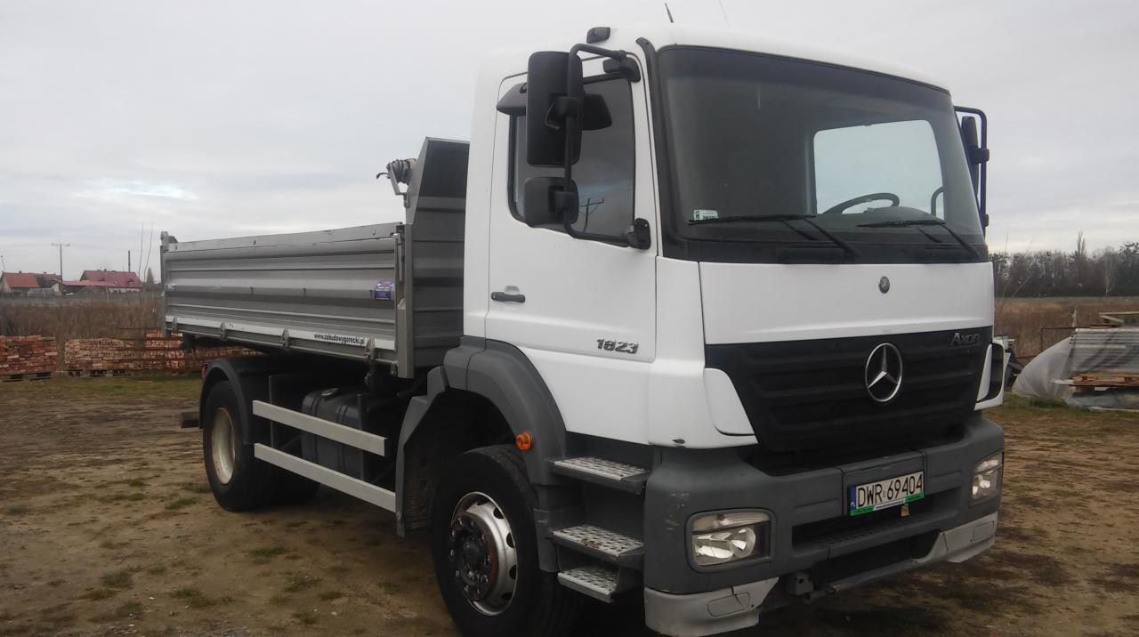 Biała wywrotka Mercedes-Benz 1823 z szarą skrzynią ładunkową na podmokłym terenie. Widoczne tablice rejestracyjne DWR 69404 i stosy drewnianych elementów w tle.