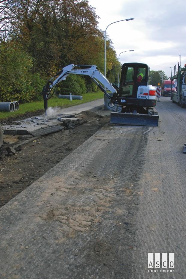 Bobcat E45 z młotem hydraulicznym rozbija starą nawierzchnię drogi, obok leżą betonowe rury, widoczne drzewa i inne pojazdy.