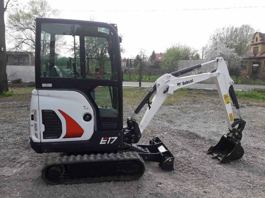 Mini koparka Bobcat E17 na żwirowym podłożu, widoczna kabina operatora, ramię koparki i łyżka, w tle budynki i drzewa.