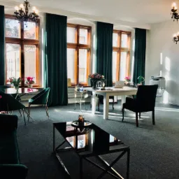 Eleganckie wnętrze biura z zielonymi akcentami: ciemnozielone zasłony, krzesła i sofa, białe biurko z laptopem, czarny stolik kawowy z lustrzanym blatem, żyrandole w stylu retro, duże okna...