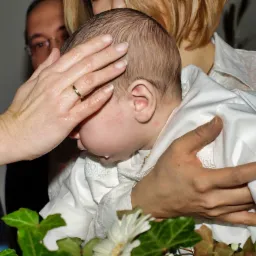 Kadr z ceremonii: dłoń z obrączką dotyka mokrej głowy niemowlęcia ubranego w białą szatkę, widoczny fragment dekoracji z bluszczu i białych kwiatów.