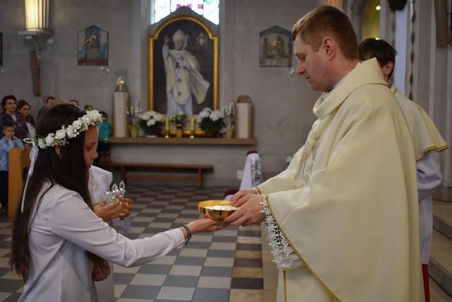 Dziewczynka w wianku komunijnym przyjmuje hostię z rąk księdza w białej szacie z haftowanymi detalami w kościele z obrazem papieża w tle.