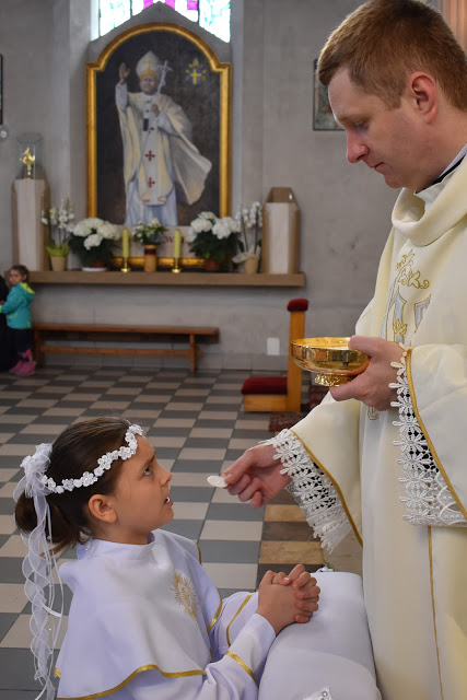Dziewczynka w białej albie i wianku przyjmuje komunię świętą od księdza w kościele, w tle obraz Jana Pawła II.