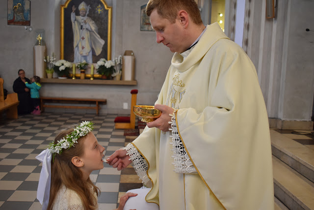 Dziewczynka w wianku komunijnym przyjmuje komunię świętą z rąk księdza w kościele, w tle obraz papieża Jana Pawła II.