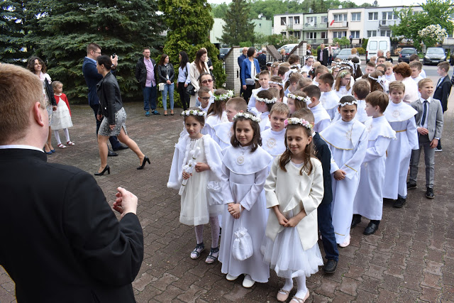 Grupa dzieci w białych strojach komunijnych stoi w rzędzie na zewnątrz budynku, prawdopodobnie przed kościołem, w tle widoczni dorośli i budynki mieszkalne.