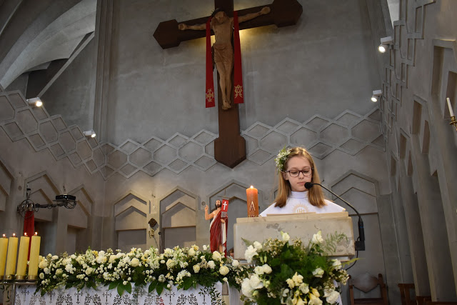 Dziewczynka w białej albie z wiankiem na głowie czyta z ambony w kościele podczas uroczystości Pierwszej Komunii Świętej, widoczny krzyż i dekoracje kwiatowe