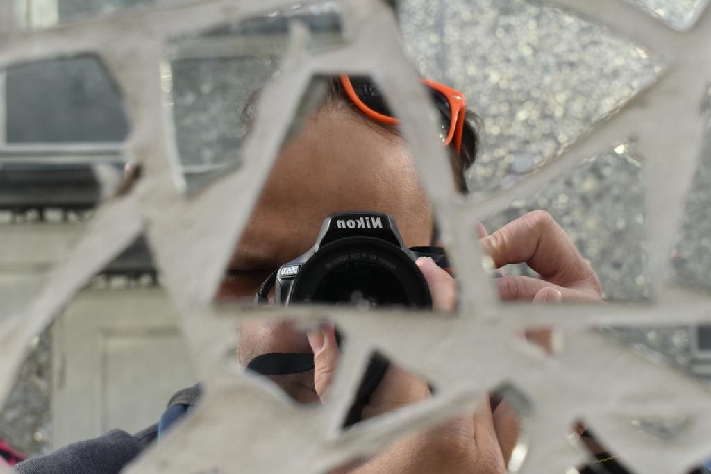 Autoportret fotografa z lustrem o nieregularnych kształtach. Widoczny fragment twarzy, aparat Nikon z obiektywem i ręce trzymające sprzęt. Pomarańczowe okulary na głowie.