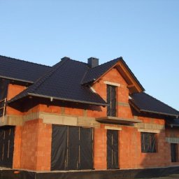 Saltan - Surowy stan domu jednorodzinnego z czerwonej cegły, z czarnym dachem i oknami zabezpieczonymi czarną folią, w trakcie budowy.