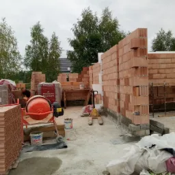 Wznoszenie ściany z czerwonych pustaków ceramicznych Porotherm, widoczna betoniarka, palety z pustakami oraz pracownik w tle, budowa w trakcie realizacji.