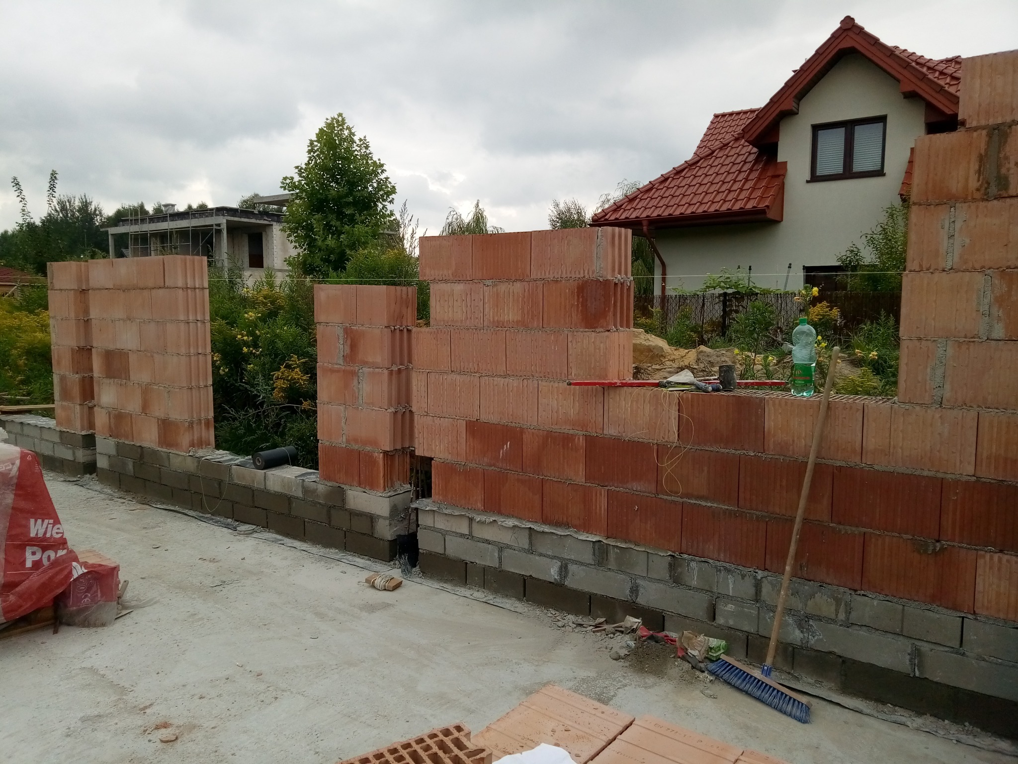 Wznoszenie muru z czerwonej cegły ceramicznej na fundamencie z szarych bloczków betonowych, widoczna zaprawa murarska, worek z materiałami budowlanymi i miotła, w tle dom z czerwoną dachówką...