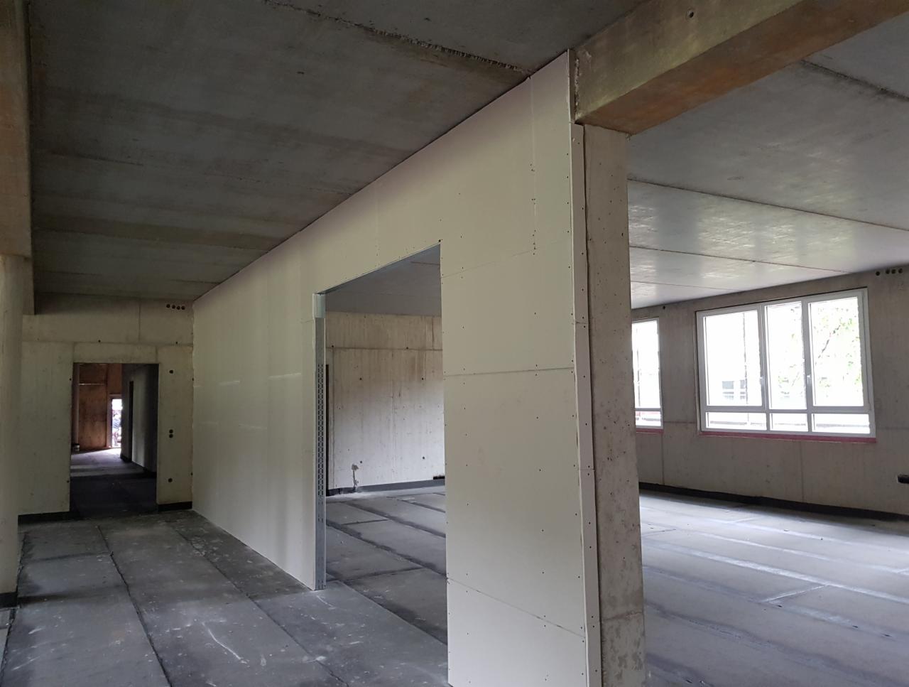 Surowy betonowy strop i ściany z płyt gipsowo-kartonowych w trakcie montażu, widoczne profile i łączenia, duże okna w tle.