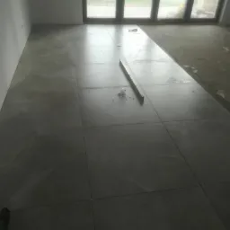 Układanie dużych, szarych płytek podłogowych w jasnym pomieszczeniu z dużym oknem; widoczna poziomica i resztki zaprawy.