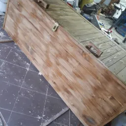 Drewniana skrzynia z metalowym zamknięciem, świeżo po lakierowaniu, widoczna na tle warsztatu z narzędziami i resztkami obróbki drewna.