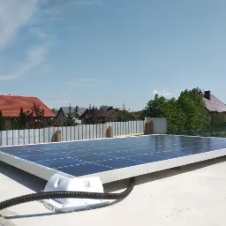 Panel fotowoltaiczny zamontowany na płaskim dachu z widocznym przewodem w peszlu, w tle zabudowa jednorodzinna i błękitne niebo.