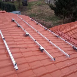 Montaż aluminiowych szyn na czerwonej dachówce, przygotowanie do instalacji paneli fotowoltaicznych, widok z góry na pochylony dach.