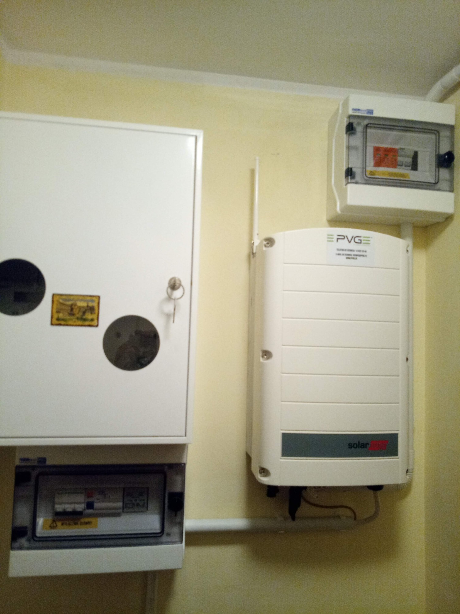 Biała szafka elektryczna z dwoma okrągłymi otworami i naklejką, obok biała skrzynka z anteną i napisem SolarEdge, oraz mała skrzynka z bezpiecznikami zamontowane na żółtej ścianie.