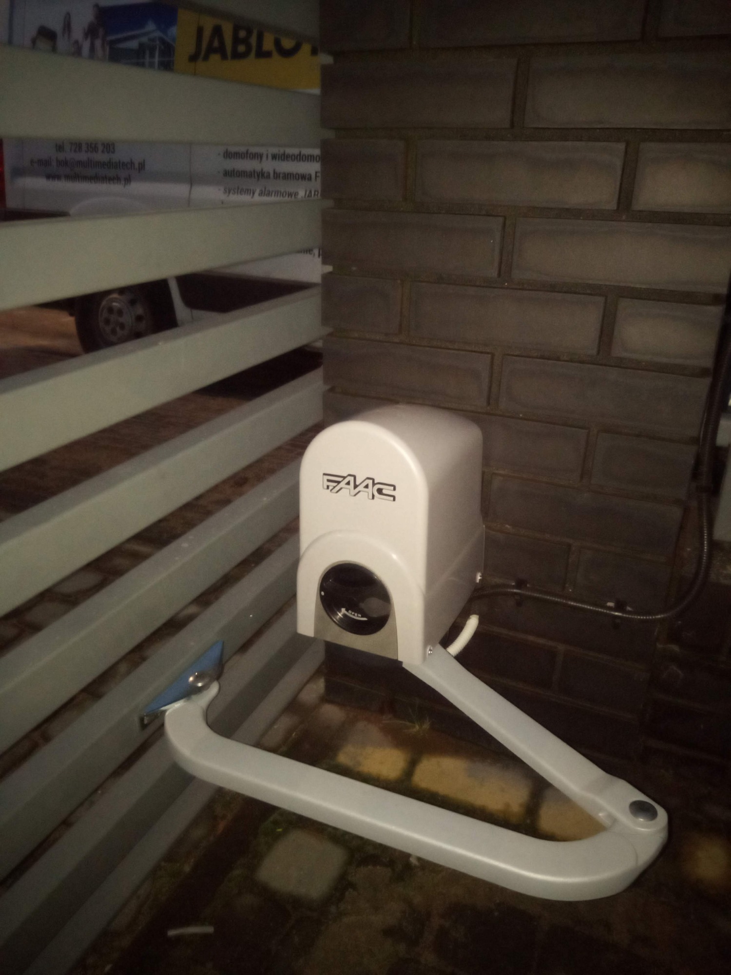 Zamontowany napęd bramy FAAC przy słupku z cegły klinkierowej, widoczna brama z metalowych profili i reklama firmy instalacyjnej w tle.