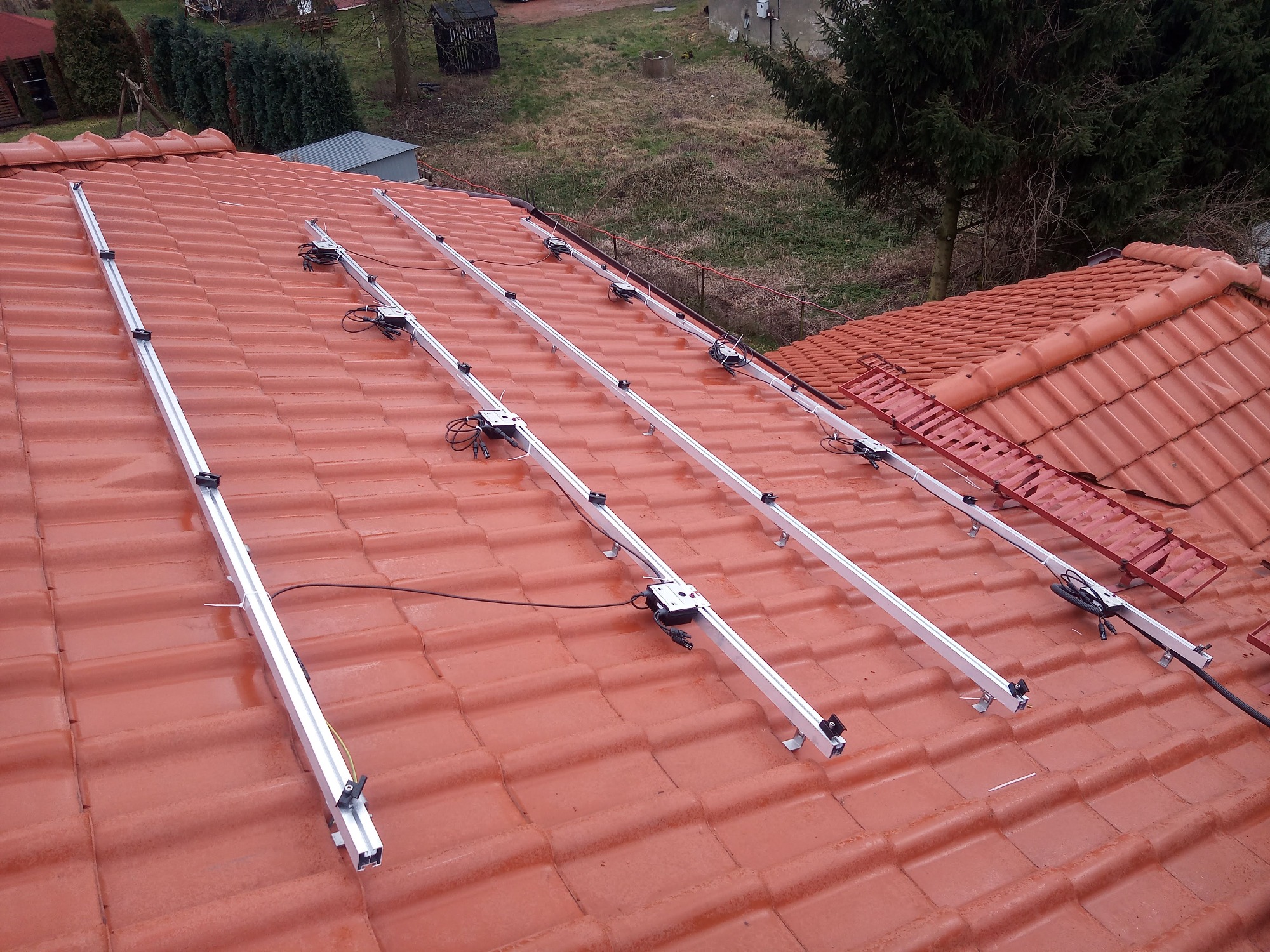 Montaż aluminiowych szyn na czerwonej dachówce, przygotowanie do instalacji paneli fotowoltaicznych, widok z góry na pochylony dach.