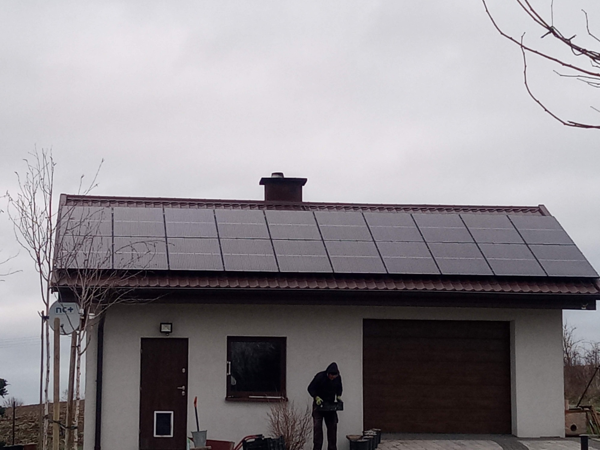 Instalacja paneli fotowoltaicznych na dachu budynku gospodarczego z brązową stolarką okienną i bramą garażową, widoczny pracownik w trakcie montażu.