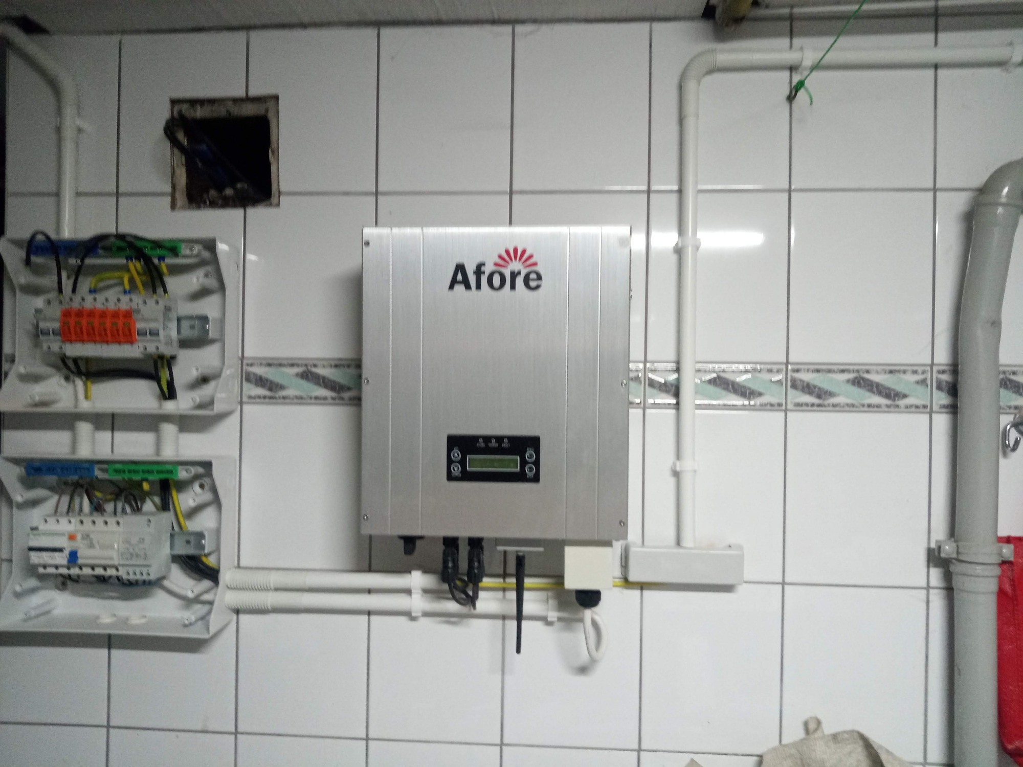 Szary inwerter solarny marki Afore zamontowany na kafelkowanej ścianie, obok otwartej skrzynki elektrycznej z widocznymi przewodami i bezpiecznikami, oraz rurami instalacyjnymi.